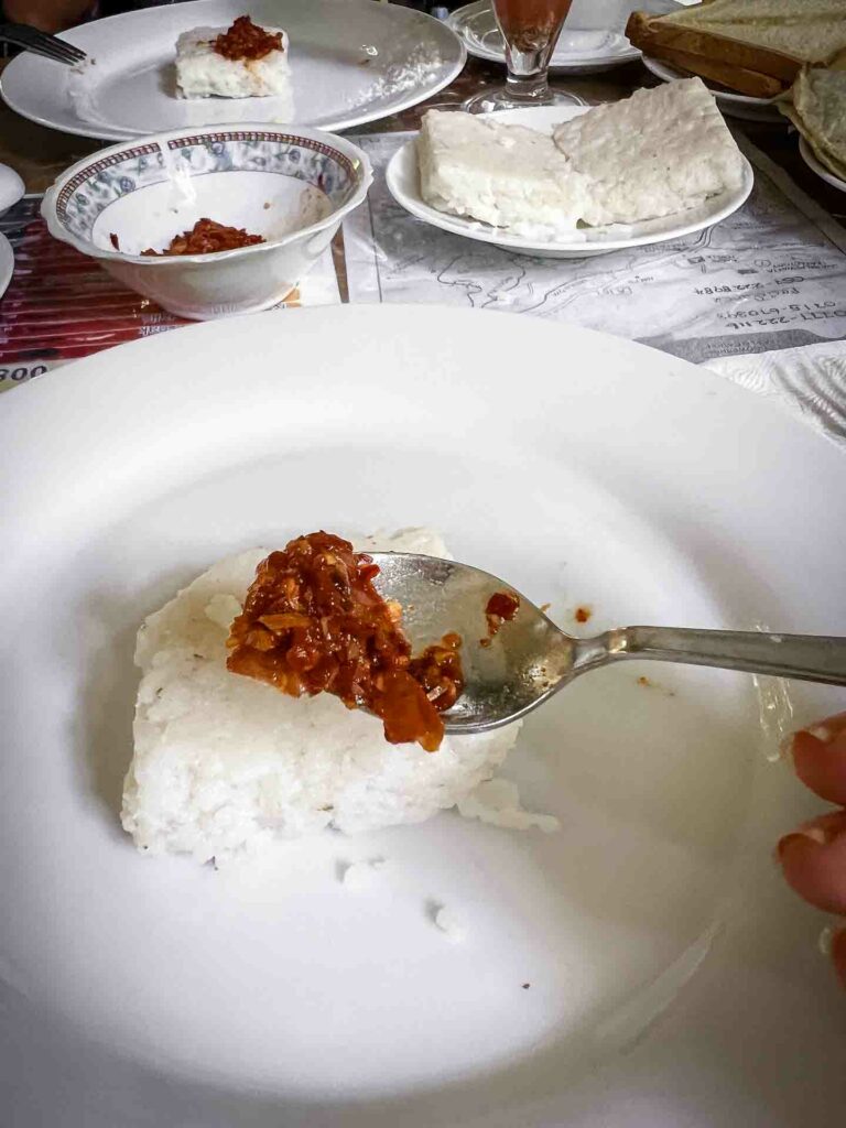 Delicious squares of Kiri Bath, a traditional Sri Lankan rice dish, neatly piled on a white plate, served alongside lunu miris sambol with chilis and maldives fish set against a rustic background in Ella, Sri Lanka.