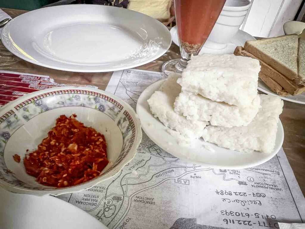 Delicious squares of Kiri Bath, a traditional Sri Lankan rice dish, neatly piled on a white plate, served alongside lunu miris sambol with chilis and maldives fish set against a rustic background in Ella, Sri Lanka.