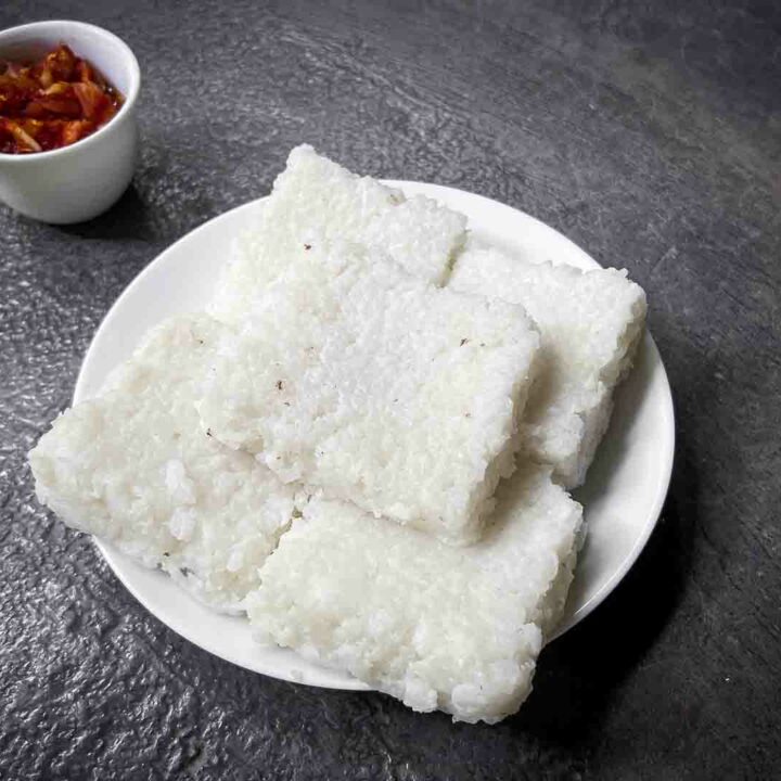 Delicious squares of Kiri Bath, a traditional Sri Lankan rice dish, neatly piled on a white plate, served alongside lunu miris sambol with chilis and maldives fish set against a rustic background in Ella, Sri Lanka.