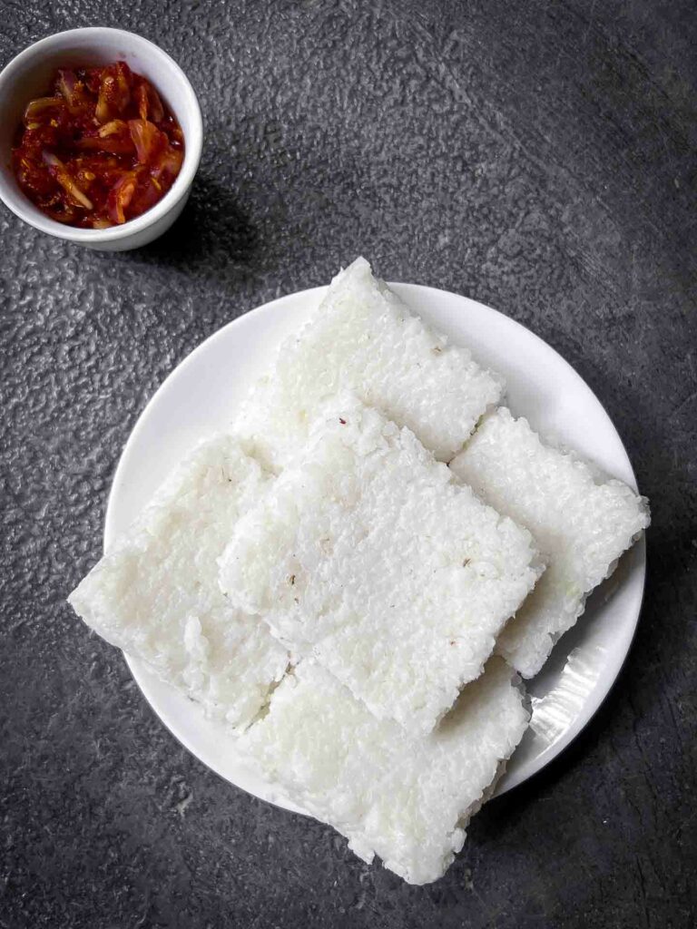 Delicious squares of Kiri Bath, a traditional Sri Lankan rice dish, neatly piled on a white plate, served alongside lunu miris sambol with chilis and maldives fish set against a rustic background in Ella, Sri Lanka.